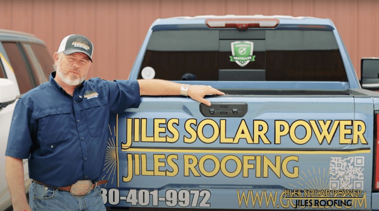 Owner of Jiles Roofing and Solar Power standing beside a branded blue pickup truck with company logo, phone number, and website displayed on the tailgate. He’s wearing a navy work shirt and branded trucker hat, with the business name prominently shown in bold yellow letters.